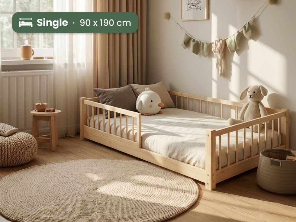 A light natural wood single toddler bed with safety rails, styled in a warm, sunlit children's bedroom with beige curtains, a woven rug, and plush toys.